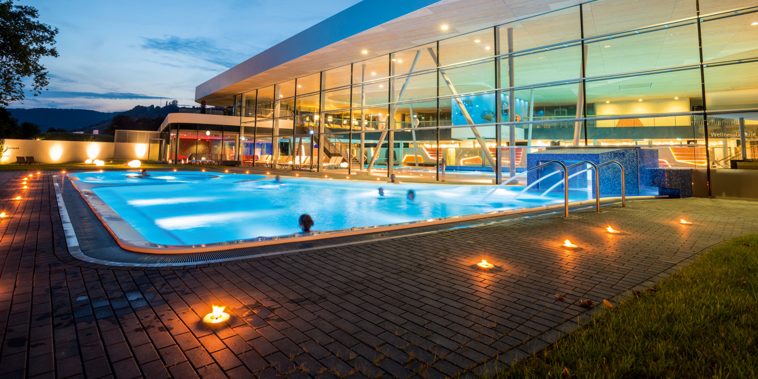 Ein modernes Schwimmbad bei Dämmerung, umgeben von einer stilvollen Gebäudeansicht mit großen Fenstern. Sanfte Beleuchtung und flackernde Kerzen schaffen eine entspannende Atmosphäre.
