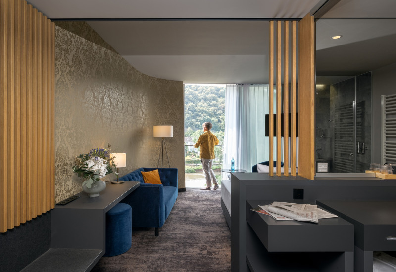 Ein elegantes Hotelzimmer mit einem blauen Sofa, einem Tisch und einer Lampe. Eine Person in gelbem Hemd steht am Fenster und blickt auf die grüne Landschaft. Einrichtungen harmonisch und modern gesta