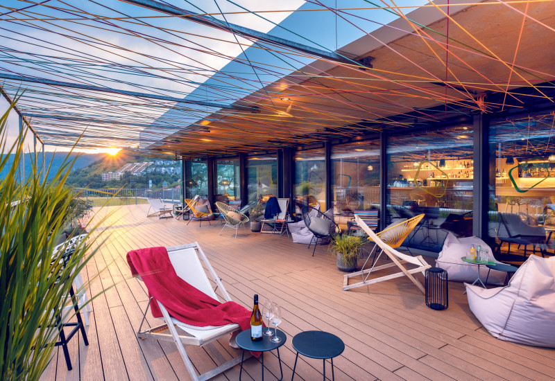 Gemütliche Terrasse mit modernem Design, geschmückt mit bunten Fäden. Sonnenuntergang im Hintergrund, Loungemöbel, Pflanzen und Getränke auf kleinen Tischen laden zum Entspannen ein.
