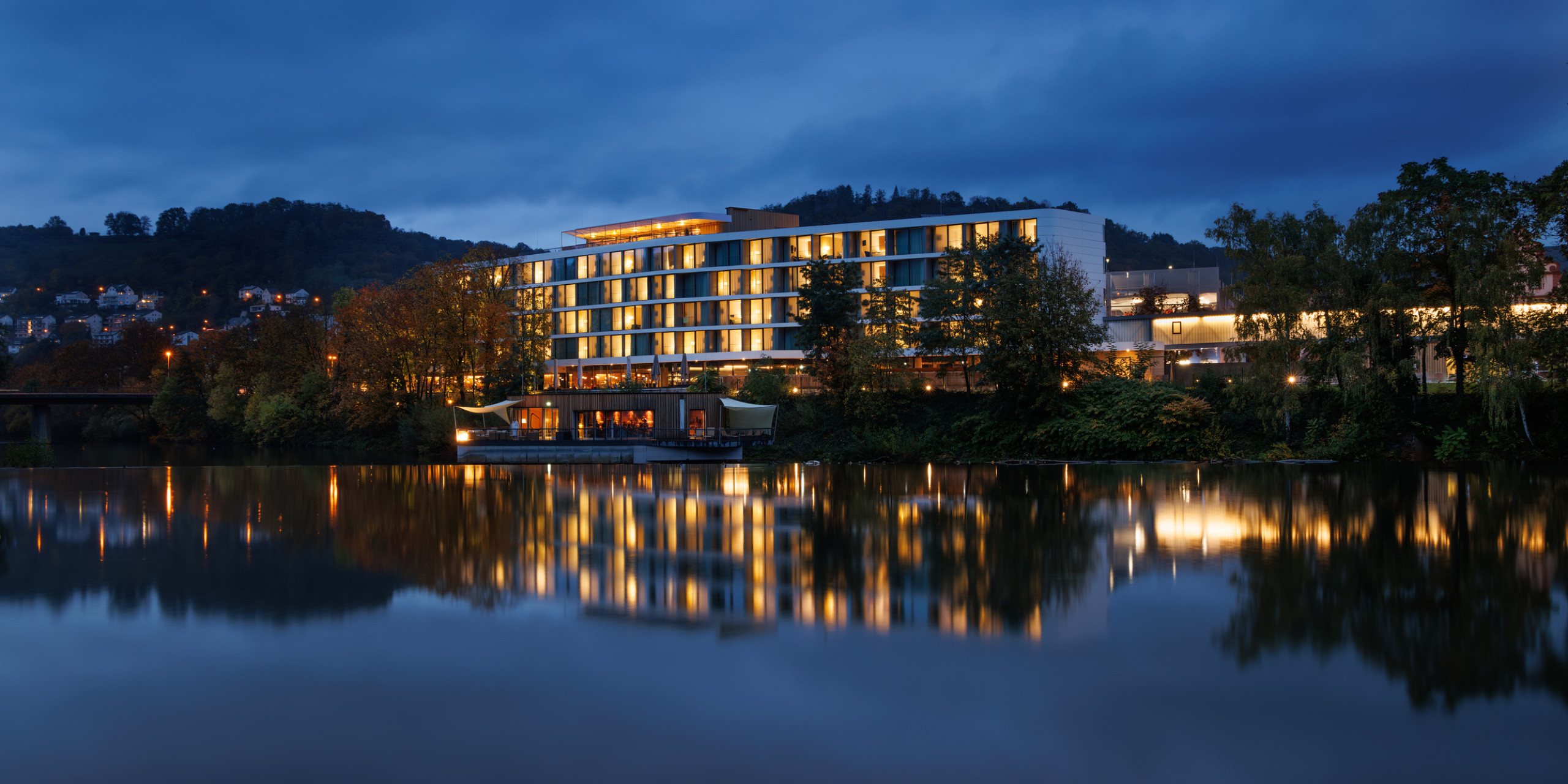 Moderne Hotelgebäude am Flussufer, beleuchtet in der Dämmerung. Bäume und Lichtreflexionen auf der Wasseroberfläche schaffen eine ruhige Atmosphäre. Sanfte Hügel im Hintergrund.