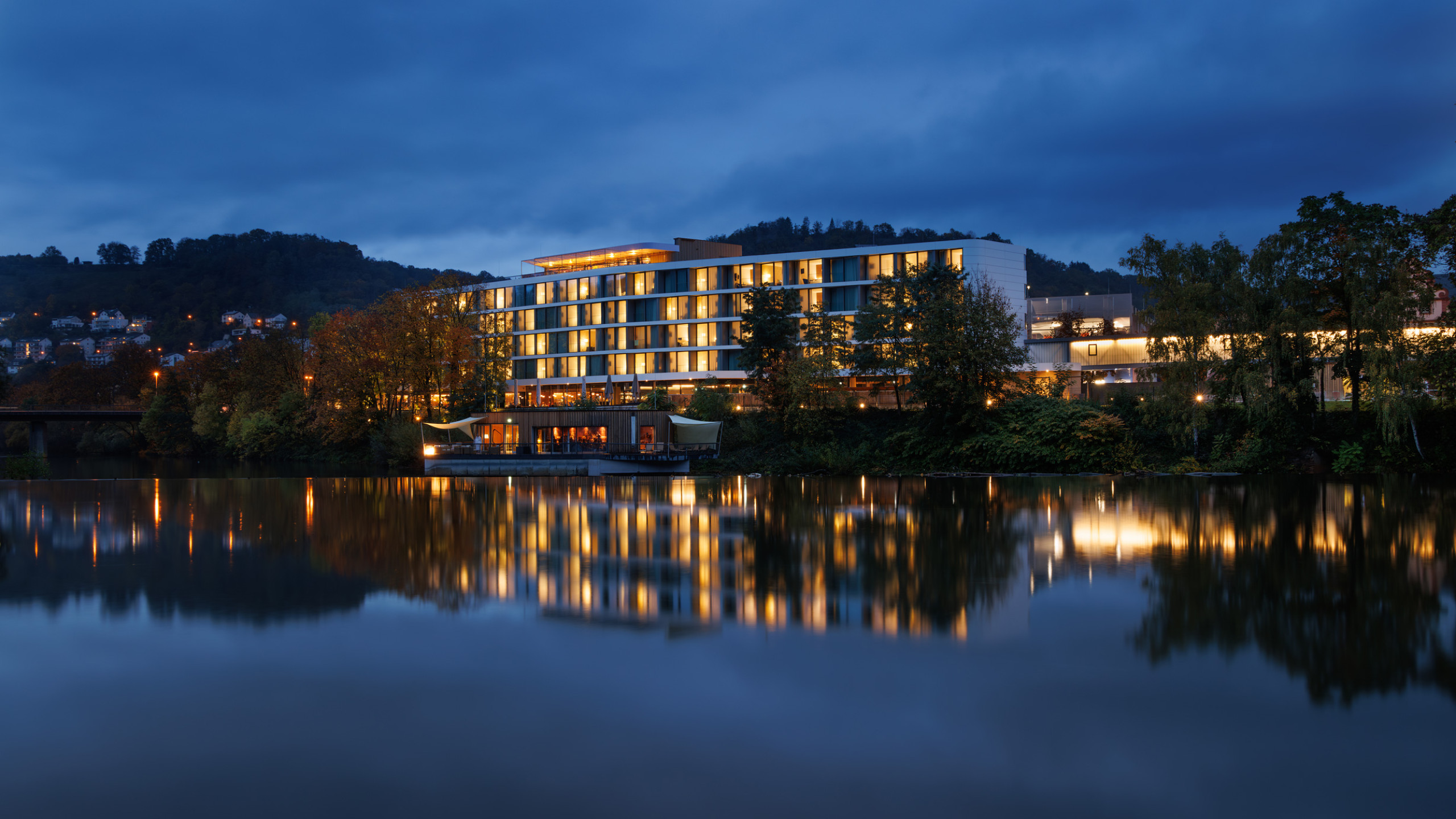 Moderne Hotelgebäude am Flussufer, beleuchtet in der Dämmerung. Bäume und Lichtreflexionen auf der Wasseroberfläche schaffen eine ruhige Atmosphäre. Sanfte Hügel im Hintergrund.