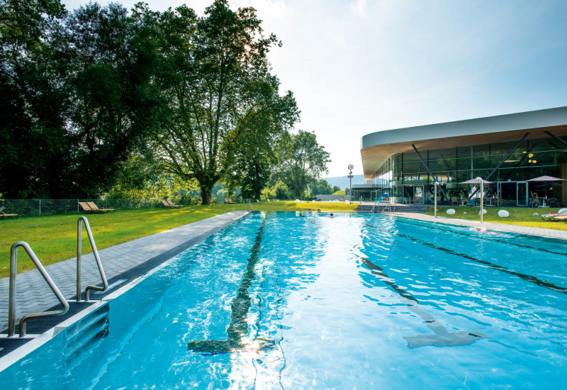 Ein einladender Schwimmbeckenbereich, umgeben von grünen Rasenflächen und Bäumen. Im Hintergrund ist ein modernes Gebäude mit großen Fenstern sichtbar, Sonnenlicht reflektiert auf dem Wasser.