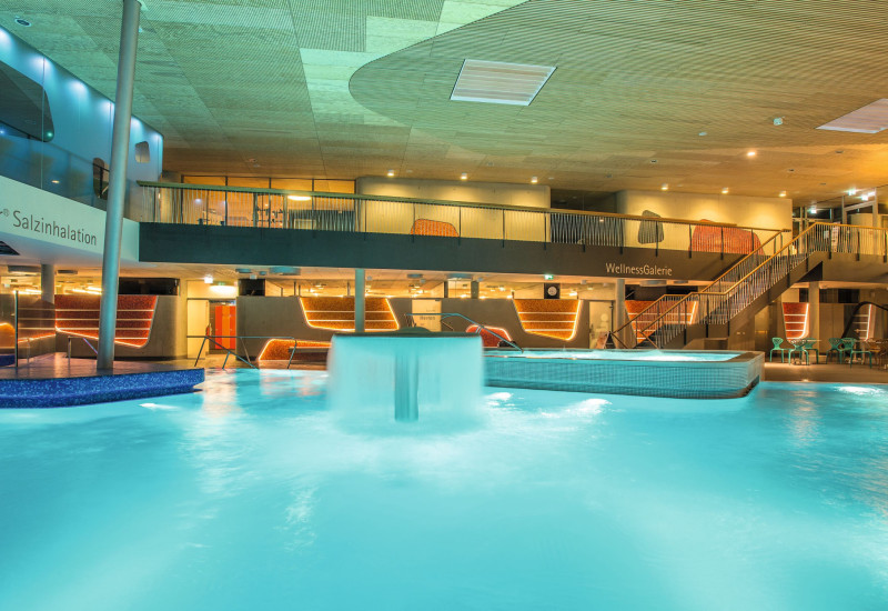 Ein modernes Wellnessbad mit leuchtend blauem Wasser. Im Hintergrund sind gemütliche Sitzgelegenheiten und stilvolle Beleuchtung zu sehen. Ein Wasserfall plätschert sanft in das Becken.