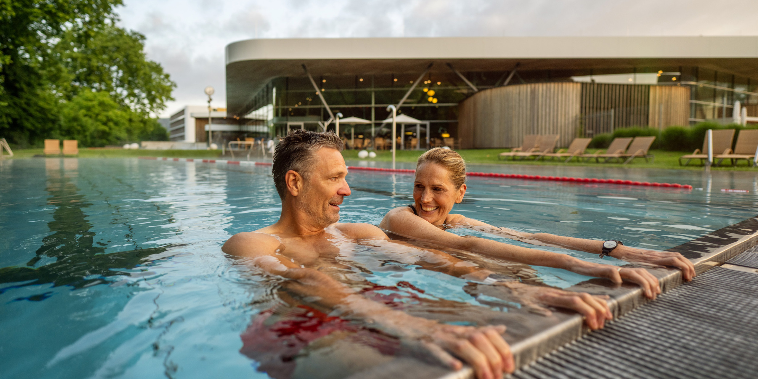 Ein Paar sitzt im Wasser eines Pools und lächelt einander an. Im Hintergrund sind Liegen und ein modernes Gebäude sichtbar, umgeben von Grün und einem bewölkten Himmel.