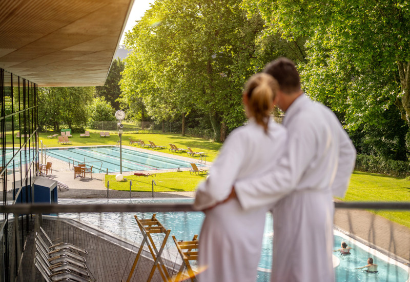 Ein Paar in weißen Bademänteln steht auf einem Balkon und blickt auf einen Außenpool, umgeben von grünem Rasen und Bäumen. Entspannte Atmosphäre mit Sonnenstrahlen.