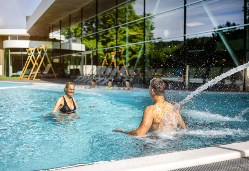 Zwei Personen genießen einen sonnigen Tag im Pool. Eine Frau in einem schwarzen Bikini lacht, während ein Mann im Wasser spritzt. Im Hintergrund sind Liegen und eine moderne Architektur zu sehen.